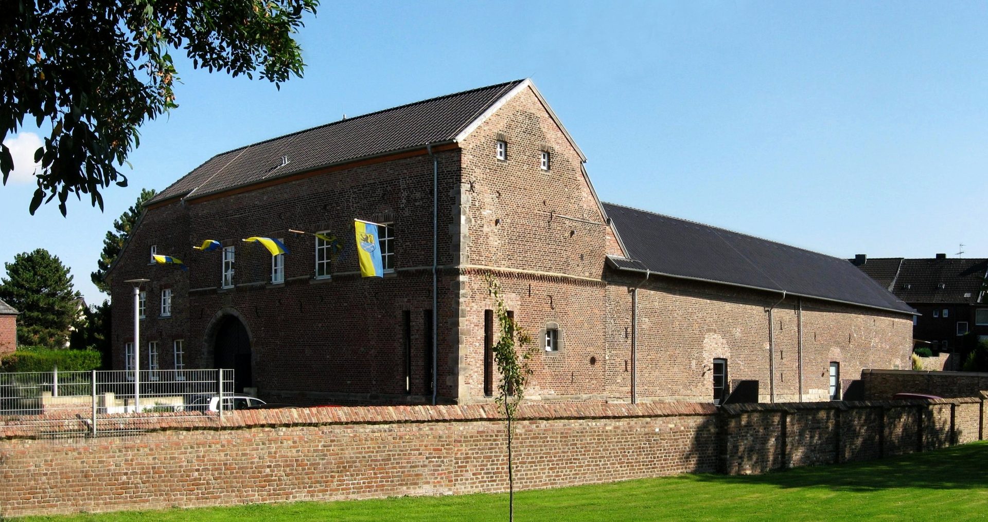 Burg Baesweiler, die backsteinerne ehemalige Wasserburg mit Torbau und Baesweiler Fahnen über der Bruchsteinmauer unter blauem Himmel