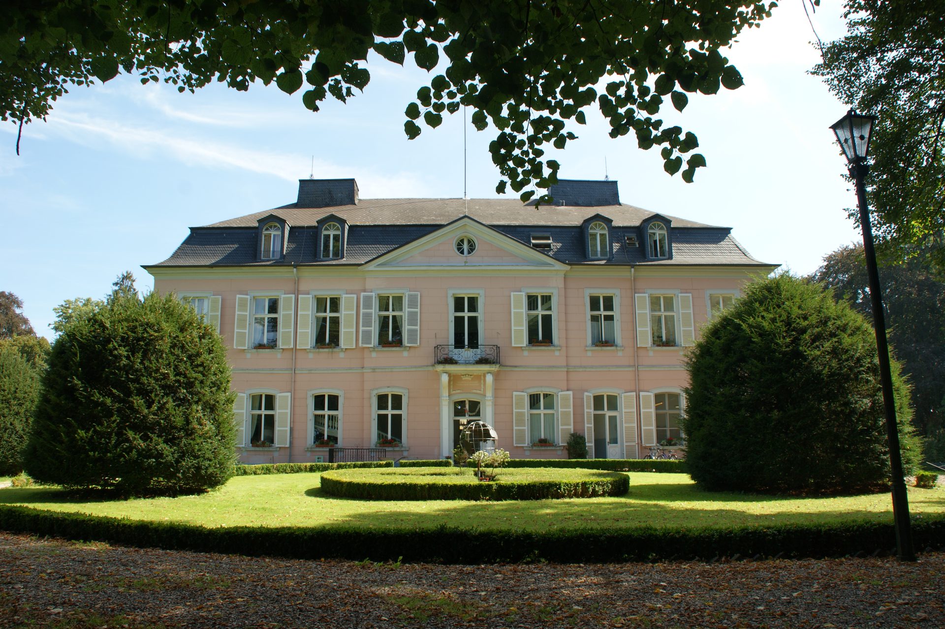 Schloss Bornheim, das rosafarbene barocke Lustschloss mit Mansarddach und Schlossgarten, gerahmt von den Zweigen eines Baumes unter blauem Himmel