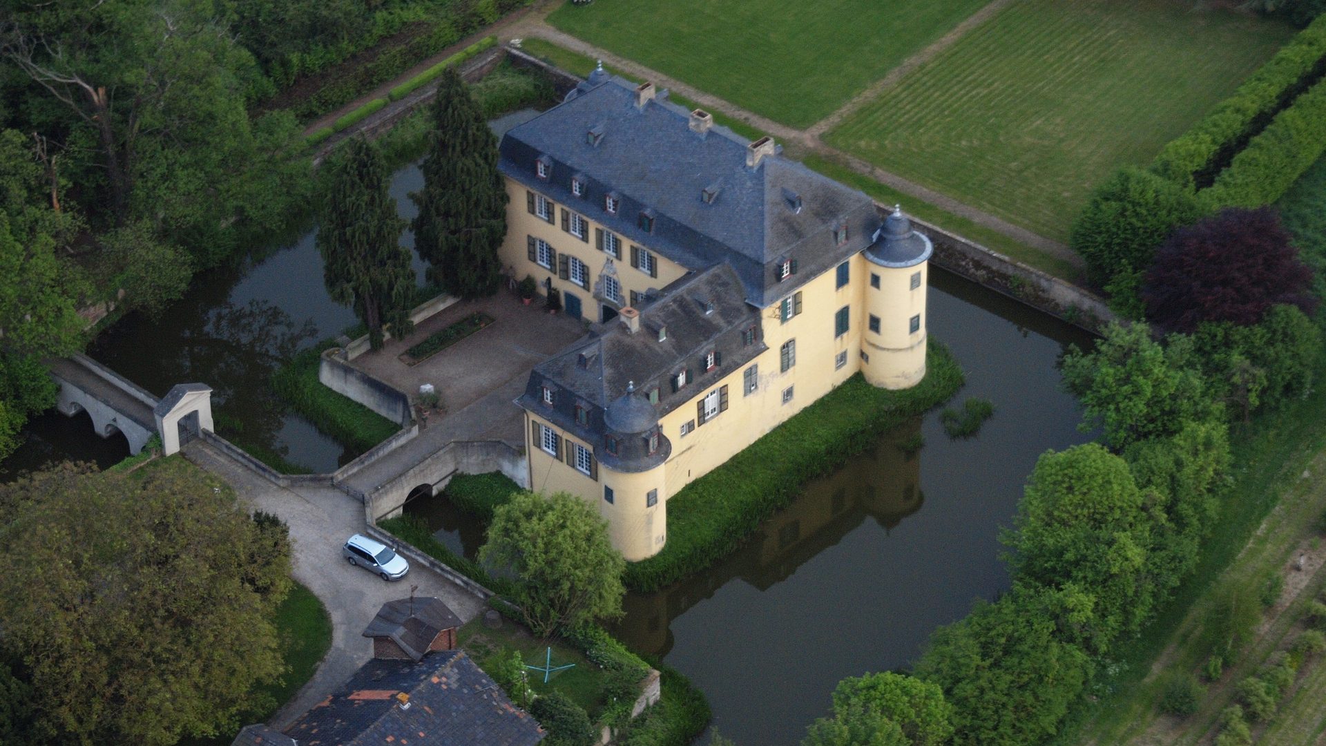 Luftaufnahme der Wasserburg Lüftelberg in Meckenheim, das barocke gelbe Wasserschloss mit Schieferdächern und Rundturm, gespiegelt im Wasser des Burggrabens und umgeben von Feldern der Voreifel